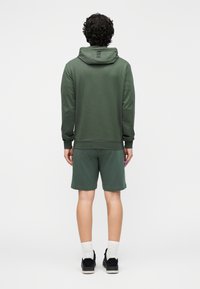 Zielona bluza z kapturem z fakturowanej tkaniny, z logo na plecach i pasujące zielone szorty. Model nosi białe skarpetki i czarne buty.