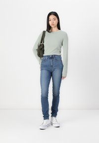 Svijetlozelena dugom rukavu, traperice visokog struka u plavoj boji i bijele platforme. Models drži maslinasto zelenu torbu preko ramena.