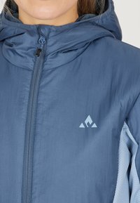 Blå lättviktsjacka med huva som har en slät yta, central dragkedja och en kontrasterande ljusblå detalj på sidan. Logotypen är placerad på bröstet.