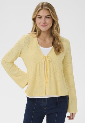 Femme aux longs cheveux blonds portant un cardigan en dentelle jaune pâle sur un haut blanc et un jean bleu foncé, debout avec une main sur la hanche.