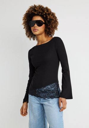 Žena s kučeravými vlasmi nosiaca veľké čierne slnečné okuliare, priliehavé čierne tričko s dlhým rukávom a čipkovaným lemom, a svetlomodré džínsy na jednoduchom pozadí.