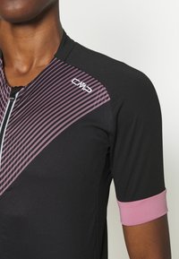 Primer plano de una persona que lleva una camiseta deportiva negra con rayas diagonales rosas y un puño de manga rosa, con un logo CMP en el pecho.