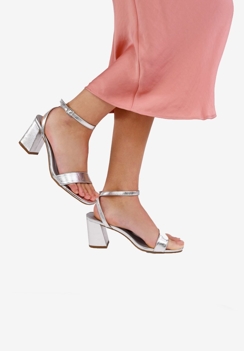 Sandali con tacco alto argentati con finitura metallica, cinturini alla caviglia, design open toe e tacco a blocco, indossati con un vestito rosa.