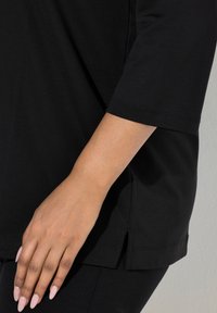 Czarny top z krótkim rękawem wykonany z miękkiej tkaniny, z bocznymi rozcięciami u dołu i luźnym krojem. Dłoń na zdjęciu spoczywa na tkaninie.