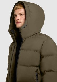 Chaqueta acolchada de color verde oliva con una gran capucha, cierre frontal con cremallera, bolsillos laterales y un diseño texturizado y acolchado que proporciona calidez y aislamiento.