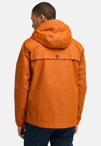 Orange vattentät jacka med huva, utrustad med elastiska ärmslut, en ryggpanel och logotypdetalj på den övre ryggen, designad för utomhusbruk.