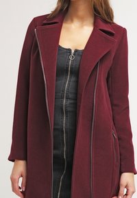 Burgundy kappa med spetsslag, asymmetrisk framdragkedja och sidofickor med dragkedja. Tillverkad av ett texturerat tyg, figurnära design.