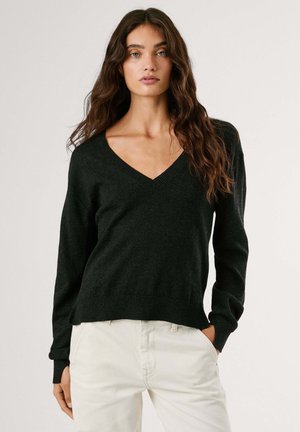 Dunkelgrüner Strick-V-Ausschnitt-Pullover mit langen Ärmeln, gerippten Bündchen und Saum, getragen über hellen Hosen. Weiche Textur, lässige Passform.