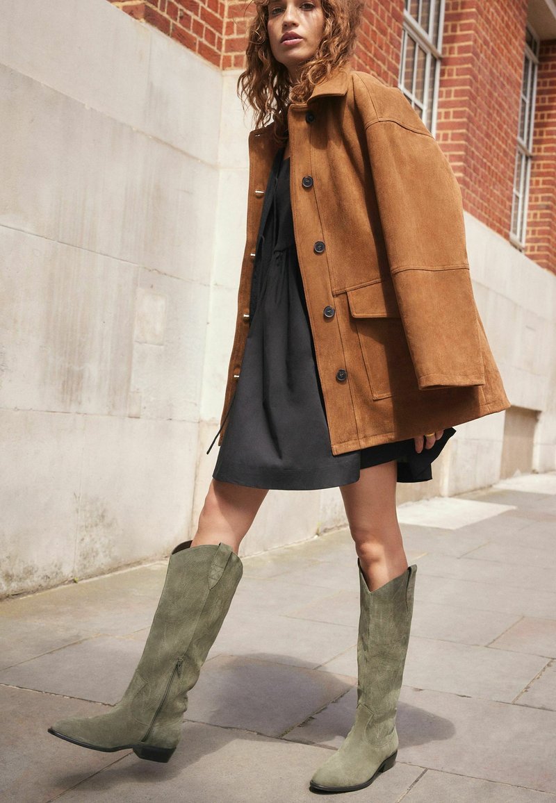 Chaqueta oversized de ante marrón con grandes botones, superpuesta a un vestido negro. Botas de ante verde hasta la rodilla con un patrón texturizado y tacón bajo.