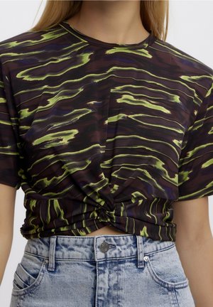 Dunkelbraunes kurzärmeliges Oberteil mit neon-grünen und violetten wellenförmigen Streifen, am Bund geknotet, getragen zu hellblauen hoch taillierten Jeans.