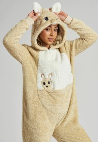 Beige fluffy onesie met capuchon en dierenoren, grote zak, zachte witte buikdetail en een kleine dierengezicht borduursel.