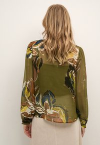 Blusa trasparente color verde oliva con motivi floreali colorati, maniche lunghe e scollatura rotonda. Il tessuto si drappeggia dolcemente, mostrando una leggera trama.