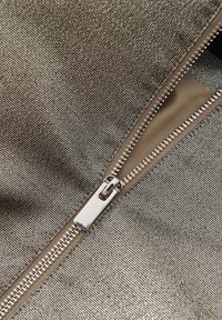 Metalen stof met een gestructureerd oppervlak, voorzien van een zilveren rits die gedeeltelijk openstaat, tegen een gladde beige voering.