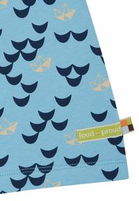Tessuto azzurro chiaro con un motivo di onde blu scuro e barche di carta color beige. Include un'etichetta verde "loud + proud" in fondo.