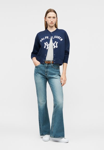 Γυναίκα που φοράει σκούρο μπλε κουμπωτό φούτερ Ralph Lauren, γκρι μπλουζάκι, καφέ ζώνη, ψηλόμεσα τζιν παντελόνια με φαρδιά μπατζάκια και άσπρα αθλητικά παπούτσια, όρθια.