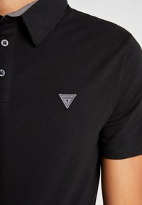 Acercamiento de una persona que lleva puesta una camiseta polo negra con un logo en forma de triángulo gris y dos botones negros en el cuello.