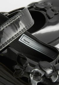 Zapatos de charol negro con punta al aire, que cuentan con una correa y un lazo decorativo. Se puede ver la etiqueta interior para personalización.