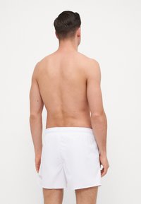 Pantaloni de baie albi, cu un brâu elastic. Material neted, lungime până la jumătatea coapsei, design minimalist fără modele sau accente vizibile.