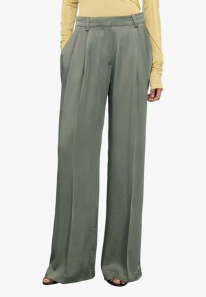 Pantaloni grigi a gamba larga con pieghe frontali, indossati con una maglia a maniche lunghe giallo chiaro infilata dentro e sandali neri con punta aperta.