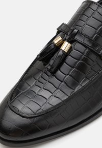 Mocasines negros de piel con brillo y estampado de cocodrilo, con herrajes en tono dorado y detalles de borlas. Textura suave con puntera redondeada.
