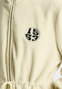 Cremefarbenes Fleece-Bademantel mit Reißverschluss, verstellbarem Bindegürtel und einem runden Logo mit den Zahlen 15 und 53 in schwarzer Stickerei.