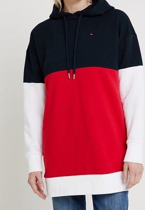 Kobieta nosząca bluzę z kapturem w blokach kolorów granatowym, czerwonym i białym, połączoną z niebieskimi dżinsami, stojąca na neutralnym tle.