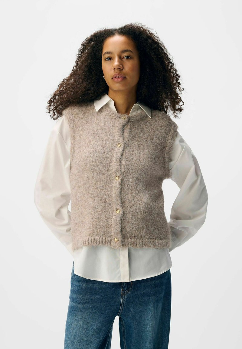 Hellbraun, flauschige Strickweste mit Knopfleiste und geripptem Saum, getragen über einem weißen Hemd mit Kragen, kombiniert mit blauen Jeans.