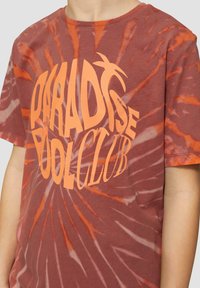 Bruine tie-dye t-shirt met een oranje spiraalpatroon en vetgedrukte tekst "PARADISE POOL CLUB." Ronde hals en korte mouwen, gemaakt van katoen.