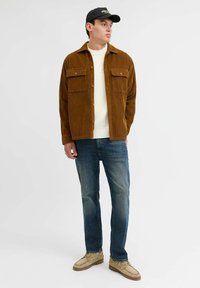 Chaqueta de camisa de pana marrón con dos bolsillos en el pecho sobre un suéter crema, combinada con jeans azules y zapatos beige, llevada con una gorra negra.