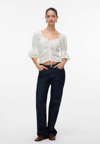 Blusa bianca, tessuto con texture, con maniche a sbuffo e scollatura quadrata, caratterizzata da un laccio in vita. Abbinata a jeans dritti di colore scuro.