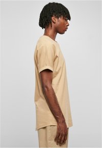 Tricou din bumbac bej cu mâneci scurte, gât rotund și o croială lejeră. Materialul are o textură netedă și o tăietură dreaptă.