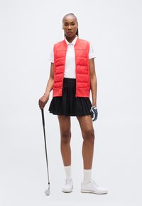 Γυναίκα που στέκεται με μπαστούνι του γκολφ στο δεξί χέρι, φοράει κόκκινο αμάνικο γιλέκο, λευκό πόλο, μαύρη πλισέ φούστα, λευκές κάλτσες και γάντι γκολφ στο αριστερό χέρι.