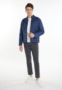 Giacca puffer blu navy con cuciture orizzontali, materiale leggero, colletto rialzato, abbinata a pantaloni grigi e sneakers bianche.