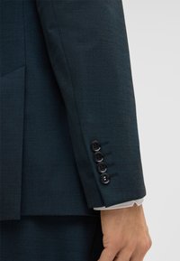 Mouw van een donker teal colbert met vier zwarte knopen, die een glimp toont van de witte shirtmanchet en een deel van een ontspannen hand.