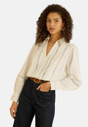 Weiße Bluse mit V-Ausschnitt, verziert mit dekorativer Spitzenkante und Puffärmeln. Kombiniert mit hoch taillierten dunklen Jeans aus Denim.