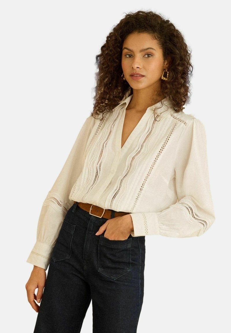 Hvid bluse med V-hals, dekoreret med blondekanter og puffede ærmer. Kombineret med højtaljede mørke denimjeans.