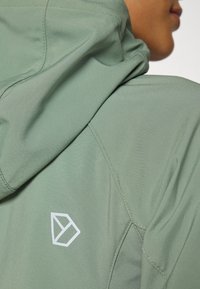 Hettejakke i lysegrønn stoff med en glatt tekstur. Har en hvit geometrisk logo på baksiden. Synlige sømmer og folder.