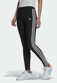 Mustat leggingsit, joissa on kolme valkoista raitaa sivuilla, ja etuosassa Adidas-logo. Yhdistetty vaaleansävyisiin lenkkareihin.