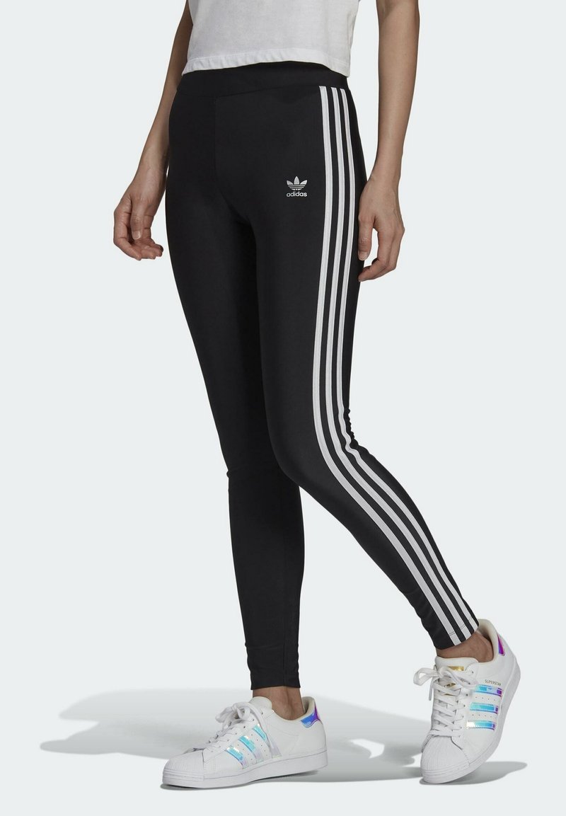 Mustat leggingsit, joissa on kolme valkoista raitaa sivuilla, ja etuosassa Adidas-logo. Yhdistetty vaaleansävyisiin lenkkareihin.