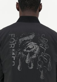 Black jacket featuring a silver graphic design on the back, depicting an eye with tears and text reading "FOR" alongside intricate patterns.