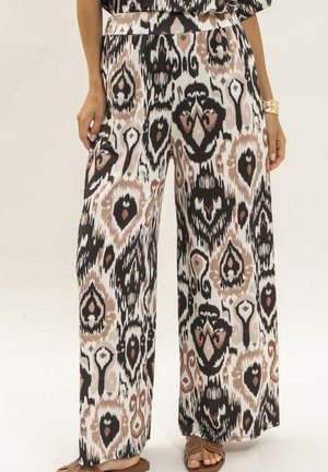 Pantalon taille large avec imprimé tribal abstrait en noir, beige et blanc, porté avec des sandales marron et un bracelet doré au poignet gauche.