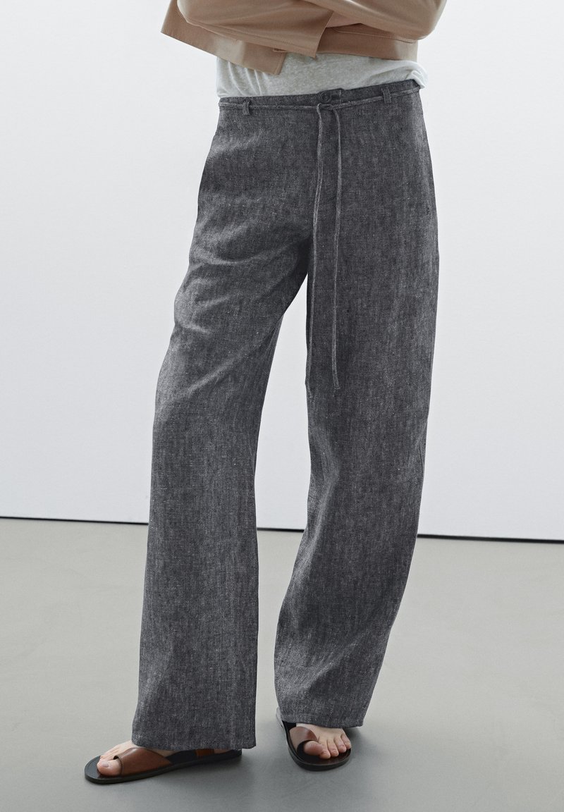 Grey, wide-leg trousers made from textured fabric, featuring a tie belt and a relaxed fit. Paired with black flat sandals.