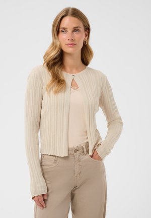 Femme aux longs cheveux blonds portant un cardigan côtelé beige par-dessus un haut clair, un pantalon beige, debout avec une main dans la poche.