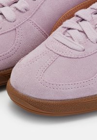 Gros plan de baskets en daim rose avec semelles en caoutchouc texturé et lacets tissés roses assortis sur fond blanc.