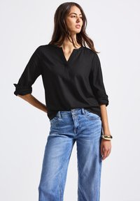 Blusa nera a maniche lunghe con colletto e polsini arrotolati, abbinata a jeans ampi di un azzurro chiaro. Accento di bracciale dorato al polso.