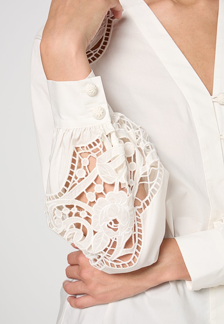 Weiße Bluse mit komplizierten Spitzenverzierungen an den Ärmeln, voluminösem Design, Knopfakzenten und einem tiefen V-Ausschnitt.