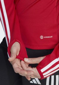 Maglietta sportiva a maniche lunghe rossa con strisce bianche, dotata di fori per il pollice. Il tessuto ha una finitura testurizzata ed è presente il logo "AEROREADY".