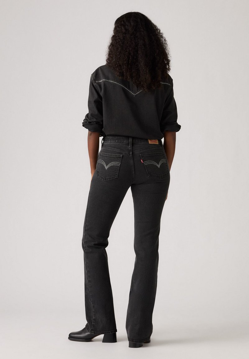 Jean en denim noir avec un design évasé, dotés de coutures contrastantes et de poches arrière caractéristiques. Associé à une chemise noire assortie.