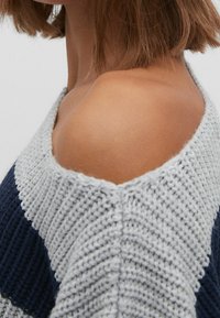 Neulottu neulepaita, jossa on harmaa ja laivastonsininen raidoitettu kuvio, leveä kaula-aukko ja teksturoitu kangas. Näkyvät olkapäät ja pehmeä, kodikas ilme.