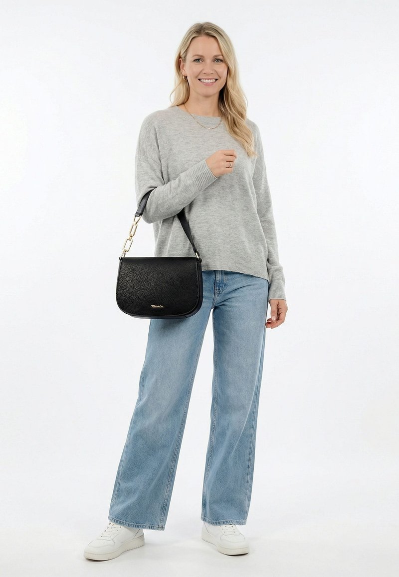 Vrouw in een grijze trui en wijde spijkerbroek, met een zwarte leren schoudertas, staand en lachend tegen een effen witte achtergrond.